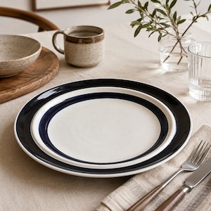 May include: A round ceramic dinner plate featuring a smooth cream-colored center and a wide, glossy dark navy blue rim. The plate has a simple, clean design with a raised edge, showcasing a high-contrast color combination of deep blue and off-white.