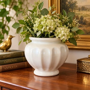 May include: A white ceramic vase with a rounded, fluted body and a wide rim. The vase is filled with a lush arrangement of green leaves and pale green hydrangea-like flowers, creating a full, rounded display of botanical elements.