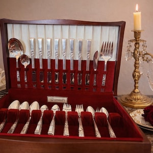 May include: A wooden silverware chest with a red velvet interior, filled with polished silver utensils. A teacup, plates, and a pearl necklace are on the left. Two lit candles and roses are on the right.