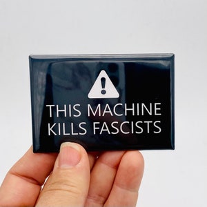 May include: A black rectangular magnet with the text "THIS MACHINE KILLS FASCISTS" in white, along with a white exclamation point symbol inside a triangle. The magnet is held in a hand.