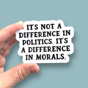 May include: White sticker with black text that reads "IT'S NOT A DIFFERENCE IN POLITICS. IT'S A DIFFERENCE IN MORALS." The sticker is held by a hand against a light blue background.