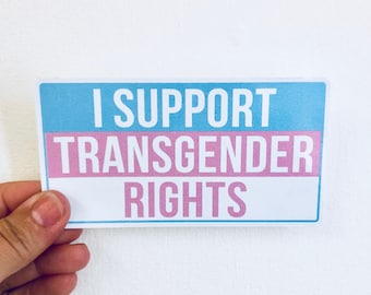 Pode incluir: Um autocolante retangular com o texto "I SUPPORT TRANSGENDER RIGHTS". O autocolante tem uma parte superior azul clara, uma secção central rosa e uma secção inferior branca. O texto é em negrito, sem serifa.