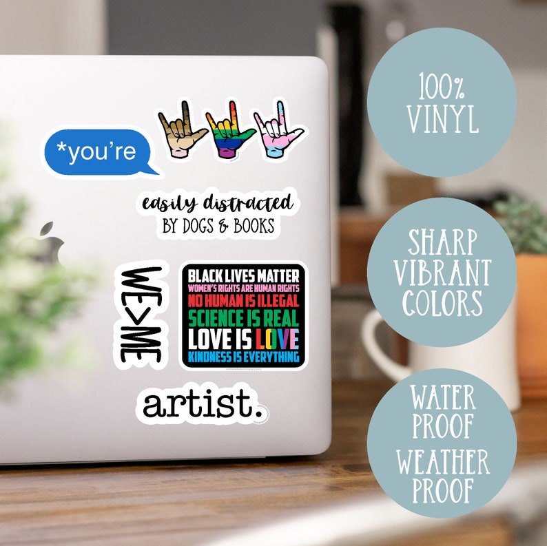 May include: A set of vinyl stickers featuring various designs, including the words "*you're easily distracted by dogs & books", "WE>ME", "BLACK LIVES MATTER", "artist.", and three hand signs. The stickers are on a laptop computer.