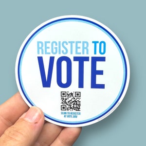 May include: A white round sticker with a blue border. The sticker says "Register to Vote" in blue text. A black and white QR code is below the text with the words "Scan to Register at Vote.gov".