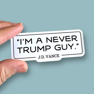 May include: A white sticker with black text that reads "I'M A NEVER TRUMP GUY." The sticker also includes the name "J.D. VANCE" below the quote. The sticker is held by a hand against a light blue background.