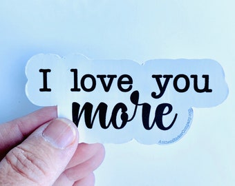 May include: White sticker with black text that reads "I love you more". The sticker is held in a hand.