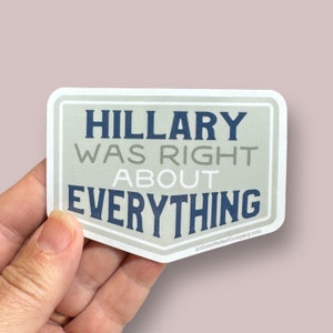 May include: A white sticker with a gray background and blue text that reads "Hillary was right about everything". The sticker is held in a hand.