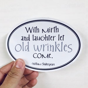 May include: Oval-shaped white sticker with black border and text. The text reads "With mirth and laughter let old wrinkles come." Below the quote is "-William Shakespeare." The sticker is held by a hand.