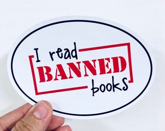 May include: White oval sticker with a black border. The sticker says "I read [BANNED] books" in black and red lettering.