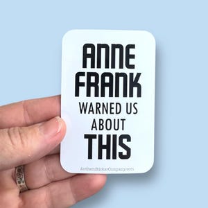 May include: A white rectangular sticker with rounded corners. Bold black text reads "ANNE FRANK WARNED US ABOUT THIS." The sticker is held by a hand against a light blue background. The website address "AnthemStickerCompany.com" is at the bottom.