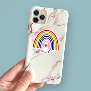 May include: A white iPhone case with a marble pattern and a rainbow sticker with a red heart in the center. The sticker says "ALLY" in black text.