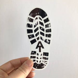 May include: Black and white sticker of a boot print with a stylized tree symbol in the center. The boot print has a distressed look.