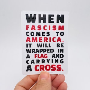 May include: A white rectangular sticker with black and red text. The text reads: "WHEN FASCISM COMES TO AMERICA, IT WILL BE WRAPPED IN A FLAG AND CARRYING A CROSS." The sticker is held by a hand.