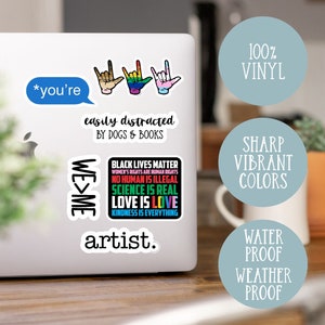 May include: A set of vinyl stickers featuring various designs, including the text "*you're easily distracted by dogs & books", "WE>ME", "BLACK LIVES MATTER", "artist.", and three hand signs. The stickers are on a laptop computer.
