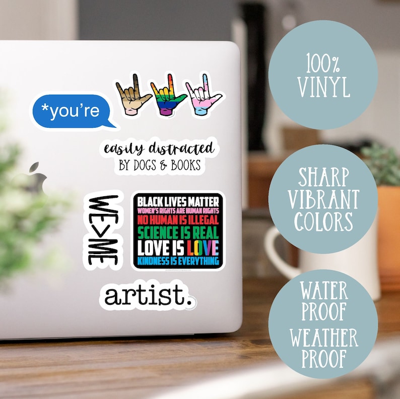 May include: A set of vinyl stickers featuring various designs, including the text "*you're easily distracted by dogs & books", "WE>ME", "BLACK LIVES MATTER", "artist.", and three hand signs. The stickers are on a laptop computer.