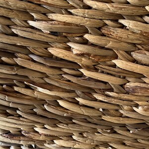May include: Close-up of a woven basket with a natural, light brown color. The basket is made of tightly woven, textured strands, creating a detailed pattern. The weave is consistent, showing the craftsmanship of the basket.