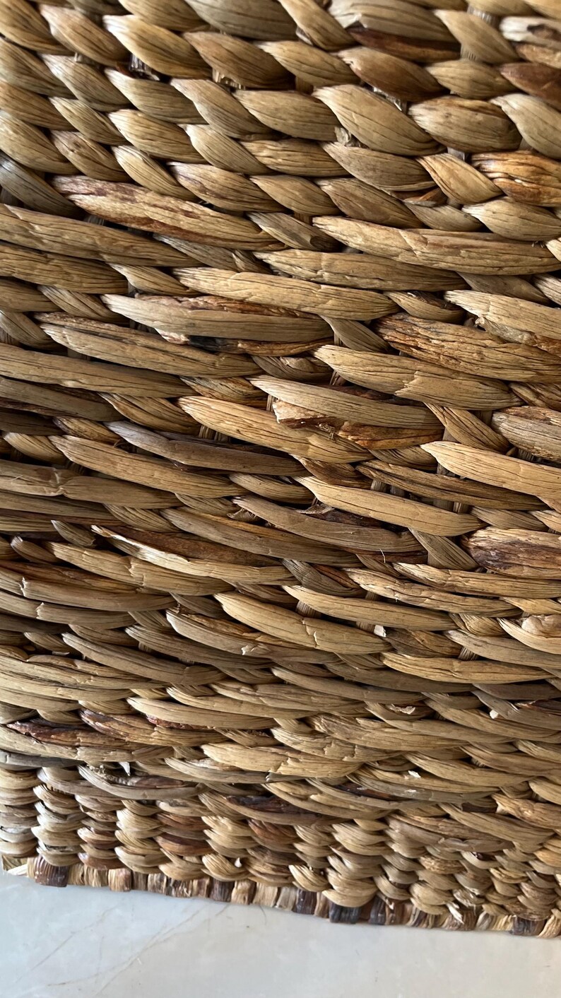 May include: Close-up of a woven basket with a natural, light brown color. The basket is made of tightly woven, textured strands, creating a detailed pattern. The weave is consistent, showing the craftsmanship of the basket.