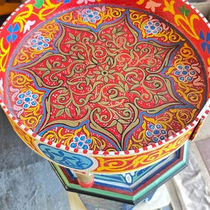 Può includere: Un vassoio rotondo dipinto a mano con uno sfondo rosso vibrante e intricati motivi floreali dorati, blu e gialli. Il vassoio è sostenuto da una base esagonale multicolore. Il diametro del vassoio è di circa 46 cm.