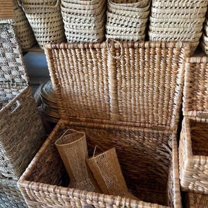 May include: A collection of woven baskets in various sizes and shapes. The baskets are made from natural-colored fibers, with a textured appearance. Some baskets are stacked, while others are open, revealing their interior. The overall aesthetic is rustic and organic.