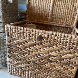 May include: Woven rectangular storage basket with a hinged lid. The basket is made of natural-colored woven material and features a wooden button closure. Another basket is visible in the background.