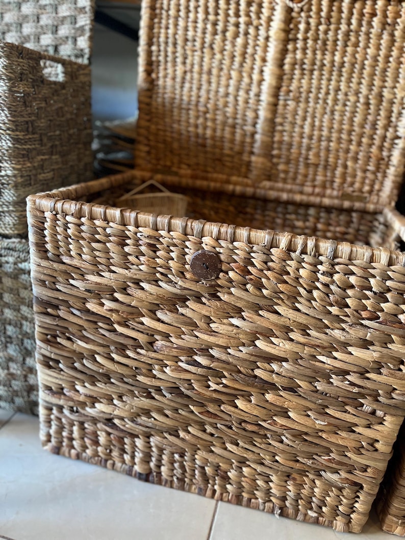 May include: Woven rectangular storage basket with a hinged lid. The basket is made of natural-colored woven material and features a wooden button closure. Another basket is visible in the background.