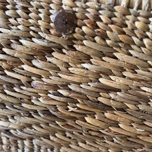 May include: Close-up of a woven basket with a natural, light brown color. The basket is made of tightly woven, textured fibers. A round, dark brown button is attached to the side of the basket, providing a contrasting accent. The basket's design suggests a rustic, handmade aesthetic.