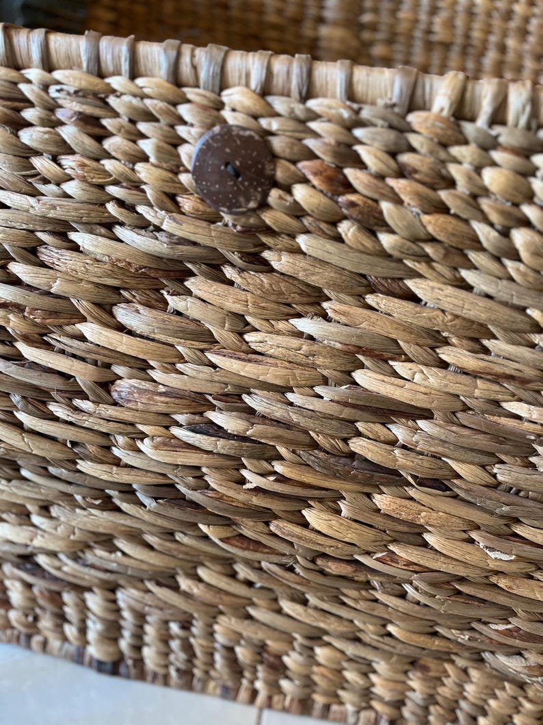 May include: Close-up of a woven basket with a natural, light brown color. The basket is made of tightly woven, textured fibers. A round, dark brown button is attached to the side of the basket, providing a contrasting accent. The basket's design suggests a rustic, handmade aesthetic.