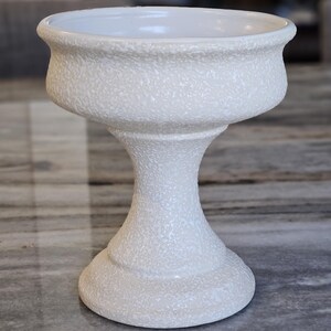 May include: A white, textured ceramic pedestal bowl with a wide, shallow top and a flared base. The bowl has a textured surface and a smooth, rounded rim. The pedestal is a decorative item, suitable for displaying small objects or as a decorative accent.