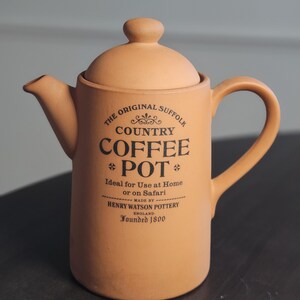 May include: A terracotta-colored coffee pot with a lid and handle. The pot has a spout and features black text that reads "THE ORIGINAL SUFFOLK COUNTRY COFFEE POT" and "HENRY WATSON POTTERY ENGLAND Founded 1800".
