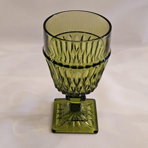 May include: A green glass goblet with a square base and a textured, patterned bowl. The goblet has a short stem and a wide, open top. The glass has a vintage look.