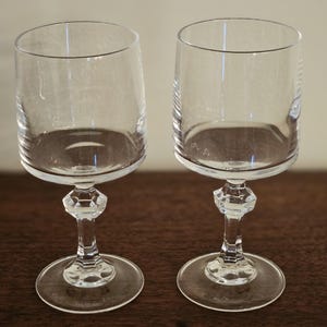 May include: Two clear crystal wine glasses with a round base, a faceted stem, and a cylindrical bowl. The glasses are displayed on a dark wooden surface. The glasses are designed for serving wine.