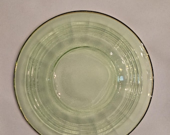 May include: A round, light green glass plate with a gold rim. The plate features concentric etched lines and a slightly raised center. The plate is likely part of a vintage or antique dinnerware set.