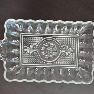 May include: Clear glass rectangular serving tray with a decorative floral and dot pattern. The tray has a scalloped edge and small, round handles on each side. The central design features a floral motif.