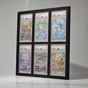 May include: A black framed display case holding six graded Pokémon trading cards. The cards feature various Pokémon characters in vibrant colors. The frame is rectangular and has a textured surface.