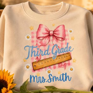Könnte beinhalten: Ein beigefarbenes Sweatshirt mit Rundhalsausschnitt und einem Grafikdruck aus einer rosa Schleife, einem Holzlineal, Gänseblümchen und gelben Sternen. Der Text Third Grade und Mrs. Smith steht in blauer Schrift auf rosa-weißem Karomuster.