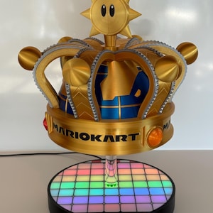 Op de afbeelding: Een goudkleurige Mario Kart kroonvormige lamp met een regenboogkleurige oplichtende basis. De kroon heeft een blauw interieur en de tekst "MARIOKART" staat erop. Een uniek decoratief item.