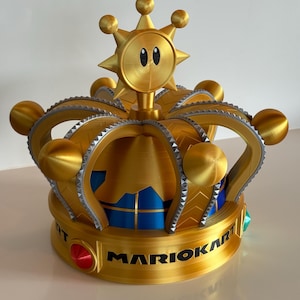 May include: A gold-coloured Mario Kart crown with a sun-like emblem on top. The crown features a detailed design with a blue and gold interior, and the words "MARIO KART" are printed on the base. The crown has a red jewel and green jewel on the base.