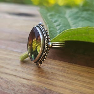 May include: A silver ring with an oval-shaped gemstone. The gemstone has a mix of colors, including green, yellow, and purple. The ring is set on a wooden surface with a green leaf in the background. The ring's band has a textured design.
