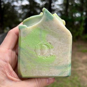 May include: A handmade bar of soap with a light green and white swirl pattern. The soap has a rounded top and a circular emblem with the letter "D" in the center. The soap is held in a hand.