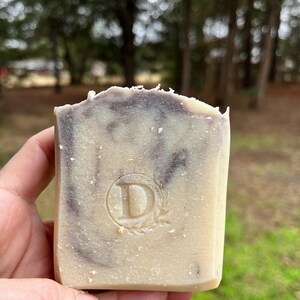 May include: A rectangular bar of handmade soap with a light beige base and dark brown swirls. The soap has a stamped "D" within a circular design. The soap is held in a hand against a blurred outdoor background.