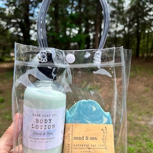 May include: A clear plastic tote bag containing a body lotion bottle and a bar of soap. The lotion bottle is light green with a white label that reads "Body Lotion, Sand & Sea". The soap is blue and white, with a brown label that reads "Sand & Sea, Handmade Bar Soap".
