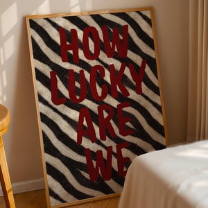 Könnte beinhalten: Ein gerahmter Kunstdruck mit einem schwarz-weißen Zebrastreifenmuster. Der Text "HOW LUCKY ARE WE" ist in kräftigem Burgunderrot geschrieben. Das Kunstwerk ist in einem hellen Holzrahmen gerahmt und an eine Wand gelehnt.