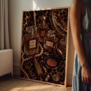 May include: A framed print featuring perfume bottles, makeup, and jewelry arranged on a leopard print background. The image includes a Coco Chanel perfume bottle, a lipstick, and a compact. The print is in a gold frame.