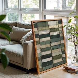 Op de afbeelding: Een ingelijst kunstwerk met een houten frame leunt tegen een beige bank. Het kunstwerk heeft een rechthoekig patroon van groene, witte en donkergroene blokken. Een glazen vaas met groene takken en een stapel boeken staan in de buurt.
