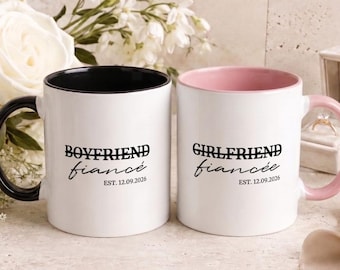 Op de afbeelding: Twee witte keramische mokken met zwarte en roze binnenkanten en oren. De linker mok heeft zwarte tekst met BOYFRIEND fiancé EST. 12.09.2026. De rechter mok heeft zwarte tekst met GIRLFRIEND fiancée EST. 12.09.2026 in een sierlijk handschrift.