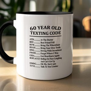 Puede incluir: Una taza de cerámica blanca con asa e interior negros, que presenta el texto impreso en negro 60 YEAR OLD TEXTING CODE seguido de una lista de acrónimos humorísticos y sus significados, incluyendo ATD, BFF, BTW, BYOT, DWI, FWIW, GHA, ROFLACGU, OMSG y TTYL.