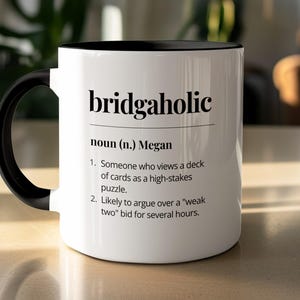 May include: White ceramic mug with a black handle and interior. The mug displays the word "bridgaholic" in black, with a definition below. The definition describes the game of bridge.