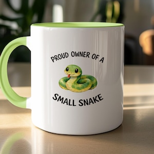 Peut inclure: Un mug en céramique blanche avec un intérieur et une anse vert clair. L'avant présente une illustration de dessin animé d'un serpent vert avec une langue rouge, entouré du texte noir "PROUD OWNER OF A SMALL SNAKE" en police sans empattement.