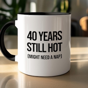 Peut inclure: Un mug en céramique blanche avec une anse et un intérieur noirs. L'avant présente un texte noir qui indique 40 YEARS STILL HOT (MIGHT NEED A NAP). Le mug a une forme cylindrique avec une finition lisse et une anse robuste sur le côté gauche.