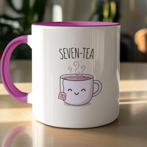 May include: A white ceramic mug with a vibrant purple handle and interior rim. The front features a graphic of a smiling tea cup with a tea bag tag labelled 70, accompanied by the text SEVEN-TEA and swirling steam lines above the cup.